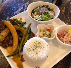 Photo: A white, square plate, containing rice, grilled pork and sausage with vegetables, and small salads