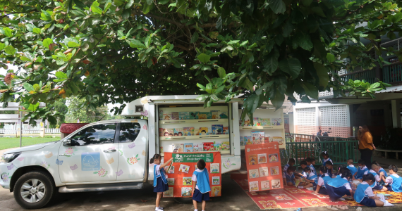A white truck painted with colorful doodles stands under a tree in front of a building. The sides of the truck are opened and reveal an assortment of children's books. A group of 15 kids in blue uniform sit behind the truck reading books. Two children stand in front of the truck, looking up at the books.