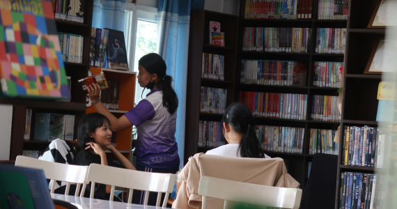 Two teenage girls are talking at a table in the library, surrounded by full bookshelves. A third girl is putting back a book into the shelf. 