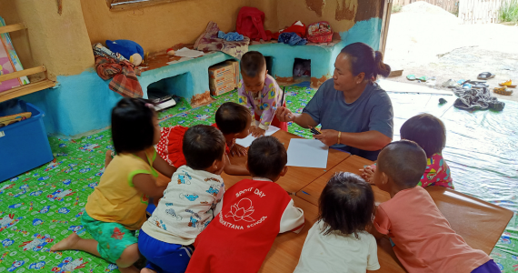 Eight children and a teacher sit around a table in a clay house. The walls are painted in a light blue. The teacher hands out paper and pencils.