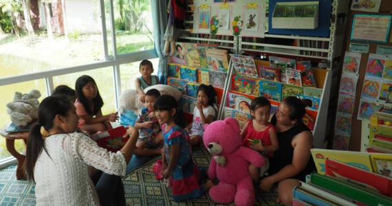 Seven kids sit in front of a bookshelf filled with big picture books. They face a librarian who is reading a story to them. There is a big window on the left and the walls are decorated with paintings.